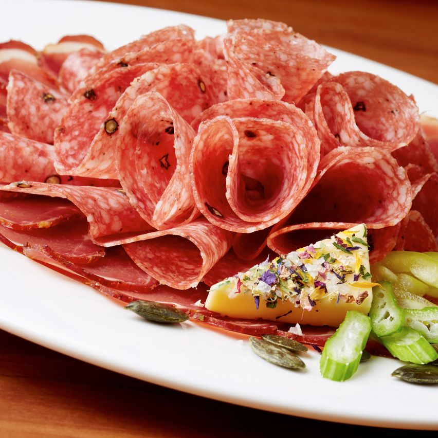 A white plate presents an assortment of cured meats, including salami, with garnishes of cucumber slices and a wedge of cheese sprinkled with herbs.