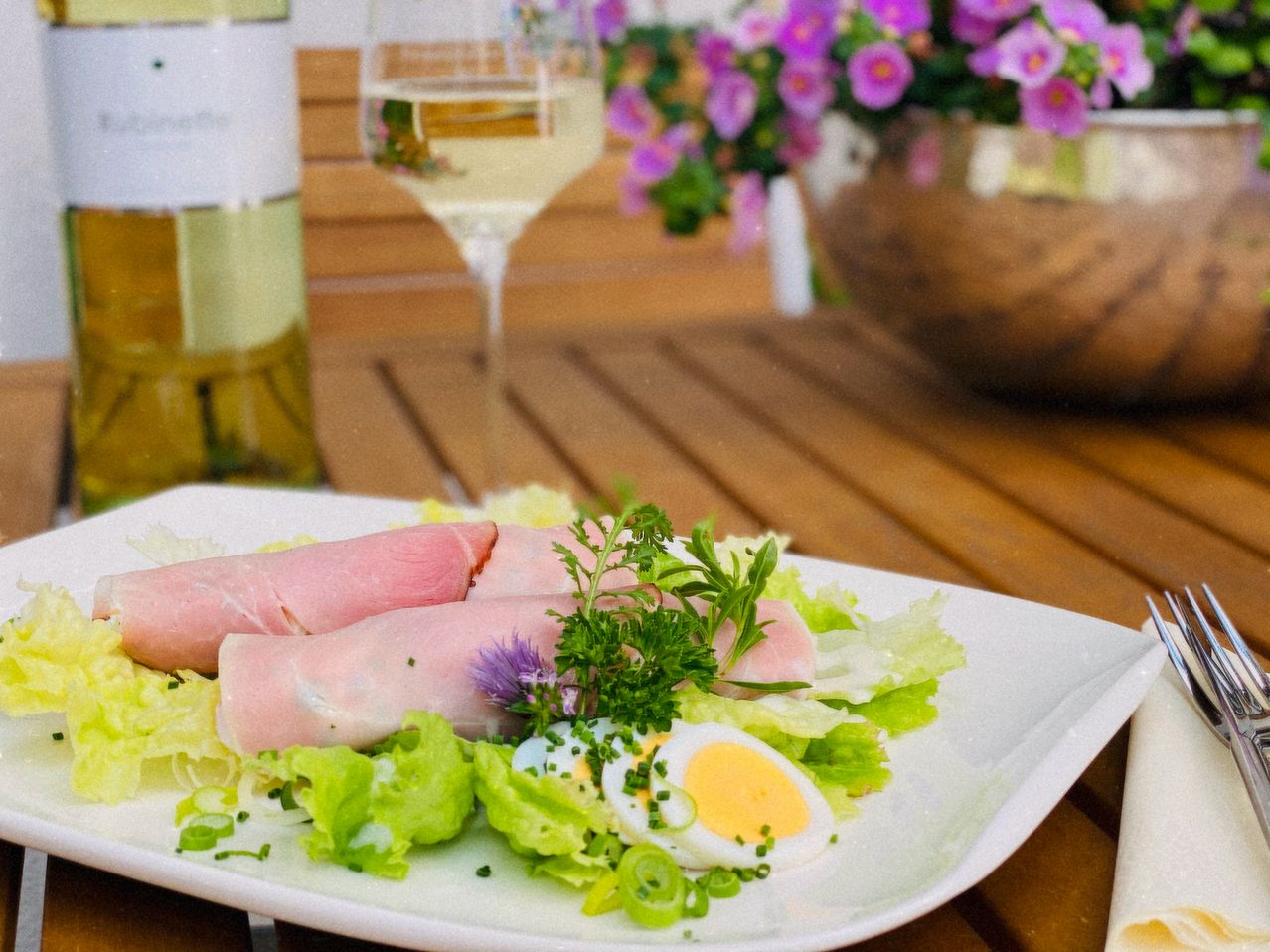 Ein Teller mit Schinkenrollen, Salat und einem gekochten Ei auf einem Holztisch, begleitet von einem Weinglas und einer Flasche Weißwein.