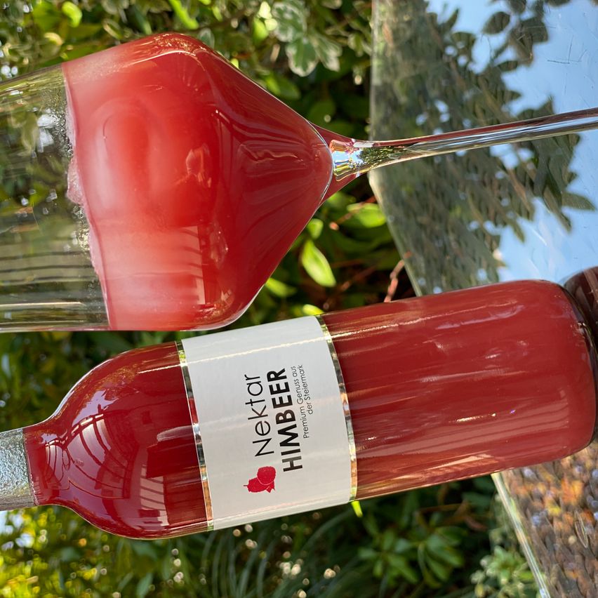 Eine Flasche Nektar Himbeer, ein rotes Getränk, steht auf einem Glastisch mit einem Weinglas, das mit demselben Getränk gefüllt ist, im Hintergrund.