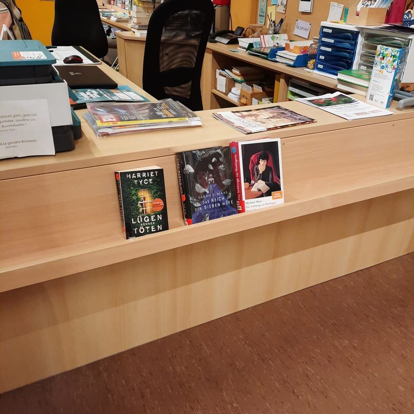 Ein hölzerner Tresen in einer Buchhandlung zeigt mehrere Bücher, darunter eines mit dem Titel 'Lugen' von Kornen Toten und ein weiteres von Harriet Tyce. Im Hintergrund befinden sich verschiedene Büromaterialien und Bücher.