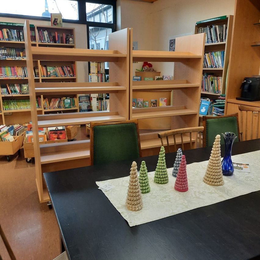 Eine Bibliothek mit mehreren Bücherregalen aus Holz, die mit Büchern gefüllt sind. Ein Tisch in der Mitte hat eine weiße Tischdecke und mehrere gehäkelte Kiefernbäume. Es gibt auch zwei Stühle mit grünen Kissen. Der Raum hat einen Holzboden und weiße Wände.