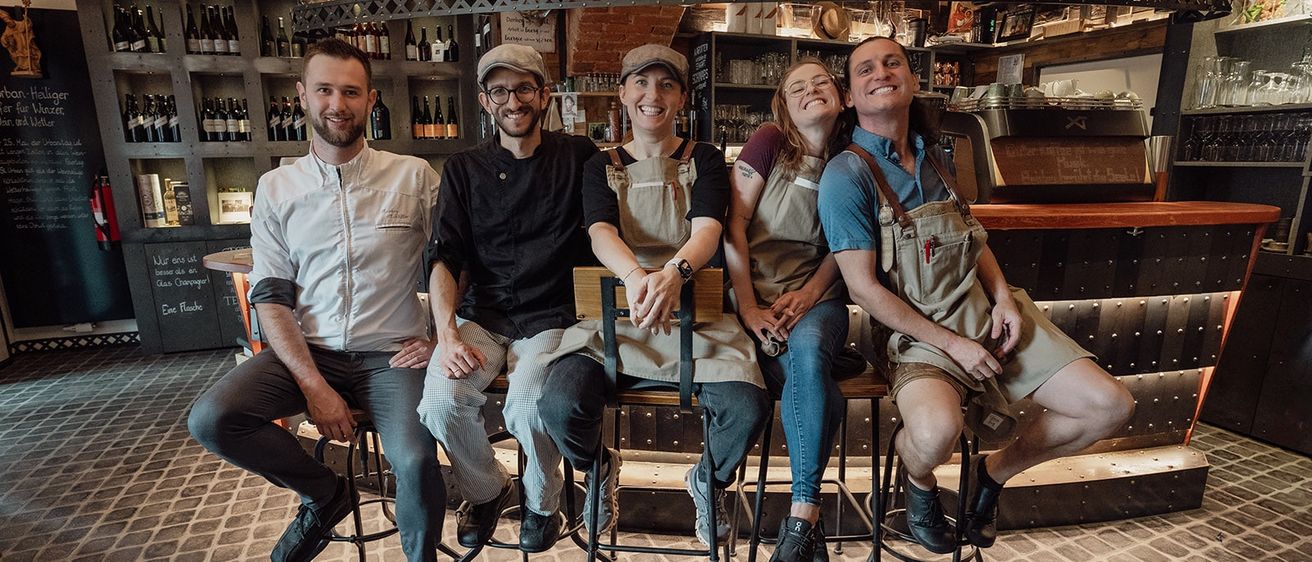 Vier Personen, zwei Männer und zwei Frauen, lächeln und posieren für ein Foto. Sie tragen Schürzen und sitzen auf Hockern vor einer Bartheke mit Regalen voller Flaschen.