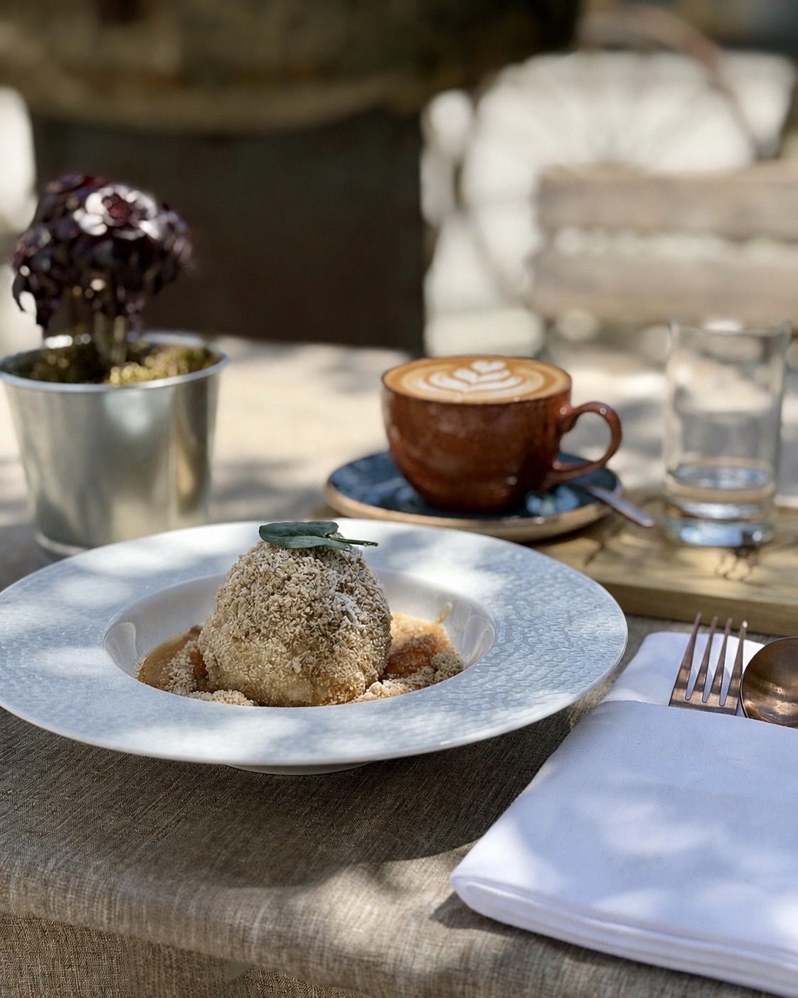 Ein weißer Teller mit einem Dessert, das mit einem Blatt gekrönt ist, steht auf einem Tisch mit einer Tasse Kaffee, einer Topfpflanze und einem Glas Wasser.