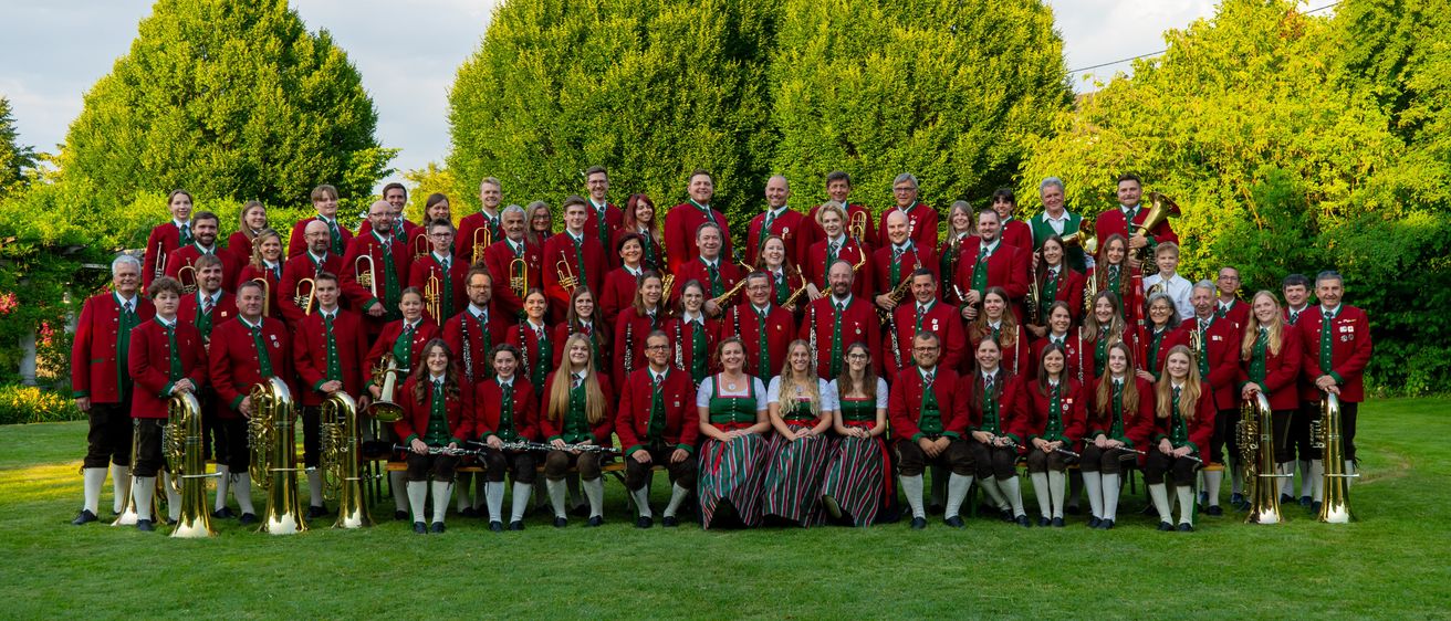 Eine Gruppe von Musikern in traditioneller Kleidung, einige halten Musikinstrumente, posiert für ein Foto auf einer Wiese mit einem grünen Busch im Hintergrund.