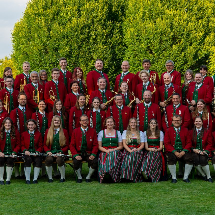 Eine Gruppe von Musikern in traditioneller Kleidung, einige halten Musikinstrumente, posiert für ein Foto auf einer Wiese mit einem grünen Busch im Hintergrund.