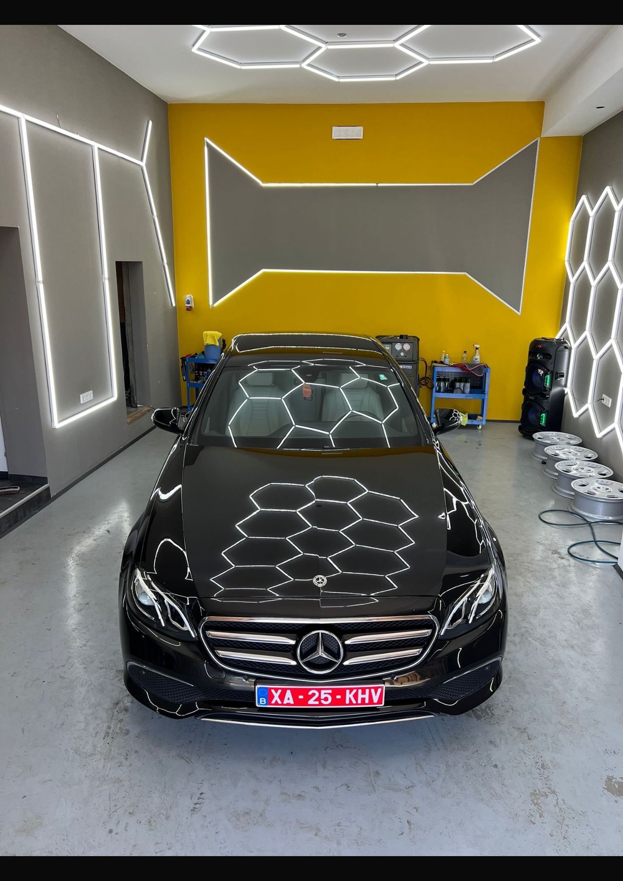 Ein schwarzer Mercedes-Benz mit einem geometrischen Muster auf der Motorhaube steht in einer Garage. Die Garage hat gelbe Wände mit sechseckigen Mustern und enthält mehrere Räder und Werkzeuge.