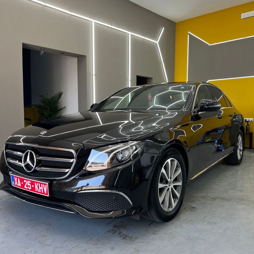 Ein schwarzer Mercedes-Benz E-Klasse Wagen mit dem Kennzeichen 'B XA-25-KHV' steht in einer Garage mit gelben und grauen Wänden, die von weißen Lichtern beleuchtet werden.
