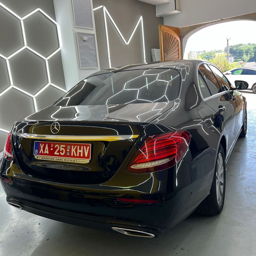 Die Rückseite eines glänzenden schwarzen Mercedes-Benz E-Klasse Autos steht in einer Garage. Das Auto hat ein rotes Nummernschild mit der Aufschrift BX 25 KHV. Die Garage hat eine weiße Wand mit einem sechseckigen Muster. Ein weißes Auto ist außerhalb der Garage geparkt.
