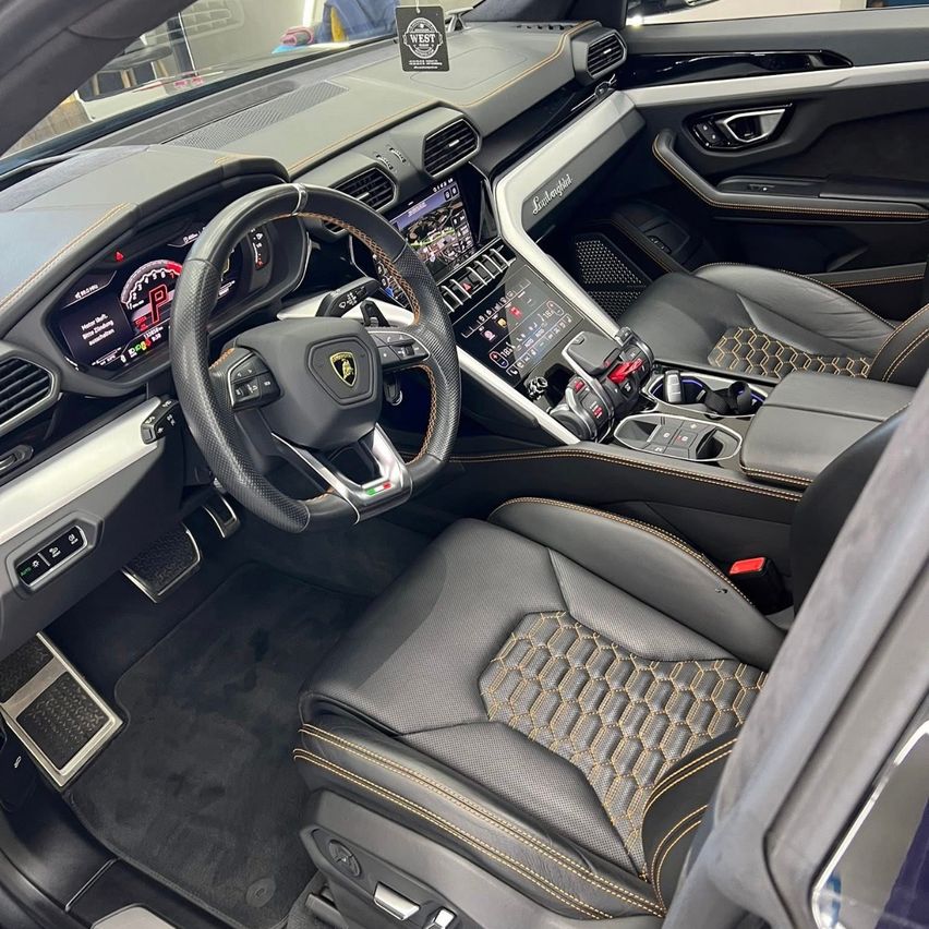 Das Interieur eines Lamborghini Urus SUV ist zu sehen. Es verfügt über schwarze Ledersitze mit goldenen Nähten und einer zentralen Konsole mit einem digitalen Display. Das Lenkrad hat silberne Akzente und einen roten Startknopf.