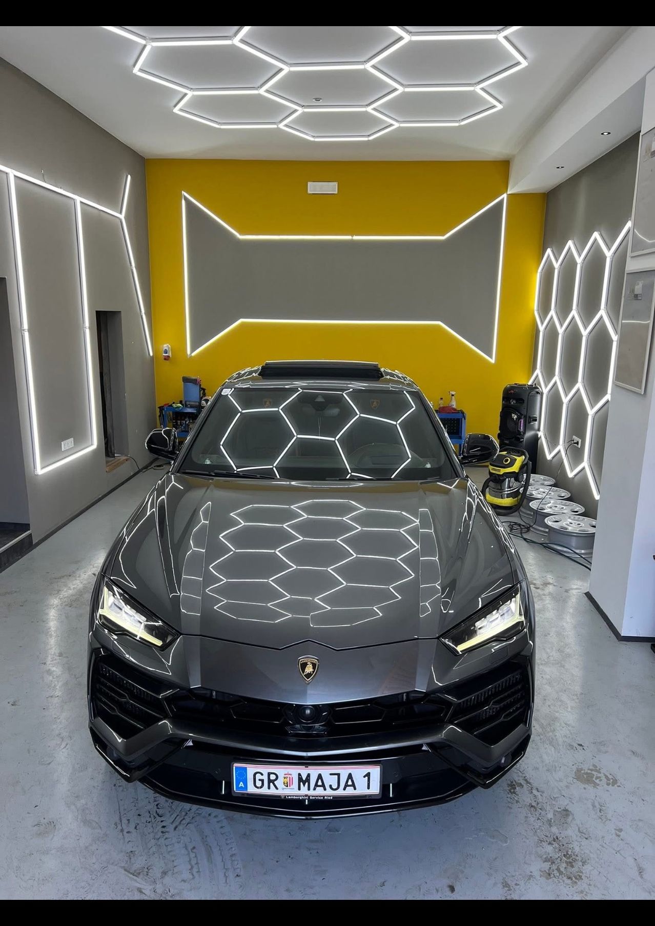 Ein grauer Lamborghini steht in einer Garage mit gelben Wänden und Sechsecklichtern. Werkzeuge und Ausrüstung sind auf dem Boden. Das Kennzeichen lautet GR MAJ1.