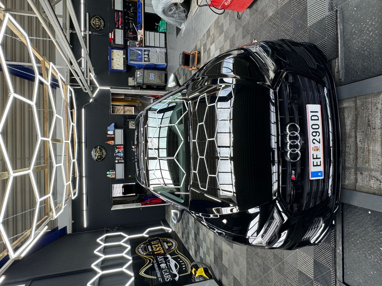 A shiny black Audi car with a hexagonal pattern on the hood is parked inside a garage. The car's license plate reads EF 2902 DL.