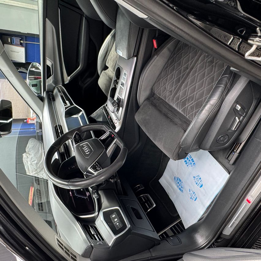 The interior of an Audi SUV shows a black steering wheel, leather seats, and a dashboard with various controls. A white bag with footprints is on the floor.