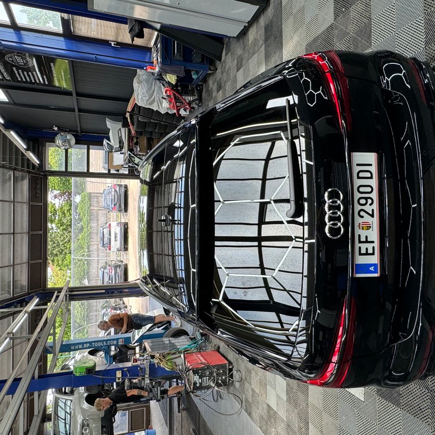 A black Audi SUV is being worked on in a garage. Two people are working on the vehicle, and it is lifted on a car lift. The license plate reads EF290DI.