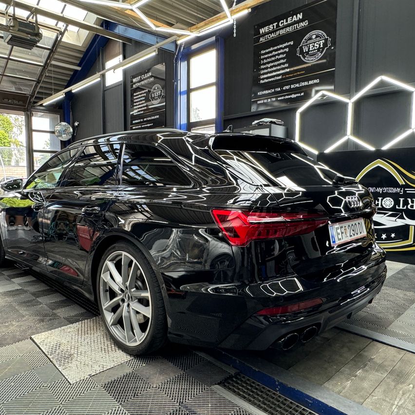 A black Audi SUV is parked inside a car wash, with its rear end visible, showcasing a polished finish and a license plate reading 'EFZ 2900D'. The car wash has a modern design with hexagonal lights and a banner with the text 'WEST CLEAN AUTOAUFBEREITUNG'.