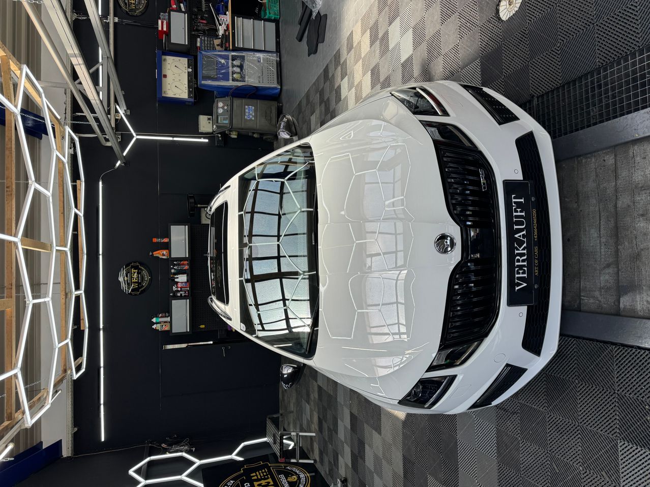 A white Skoda car is being worked on in a garage, with a checkered floor and various tools and equipment in the background.