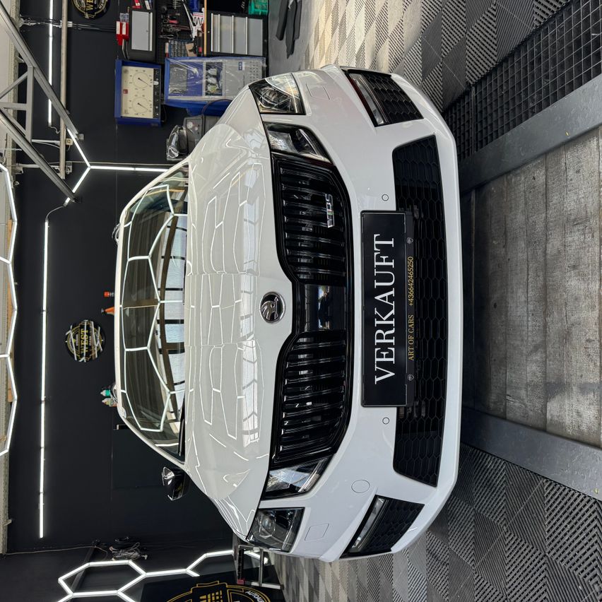 A white Skoda car is displayed in a showroom with a black and white checkered floor and a black wall. The car has a black grille with the text VERKAUFT on it.