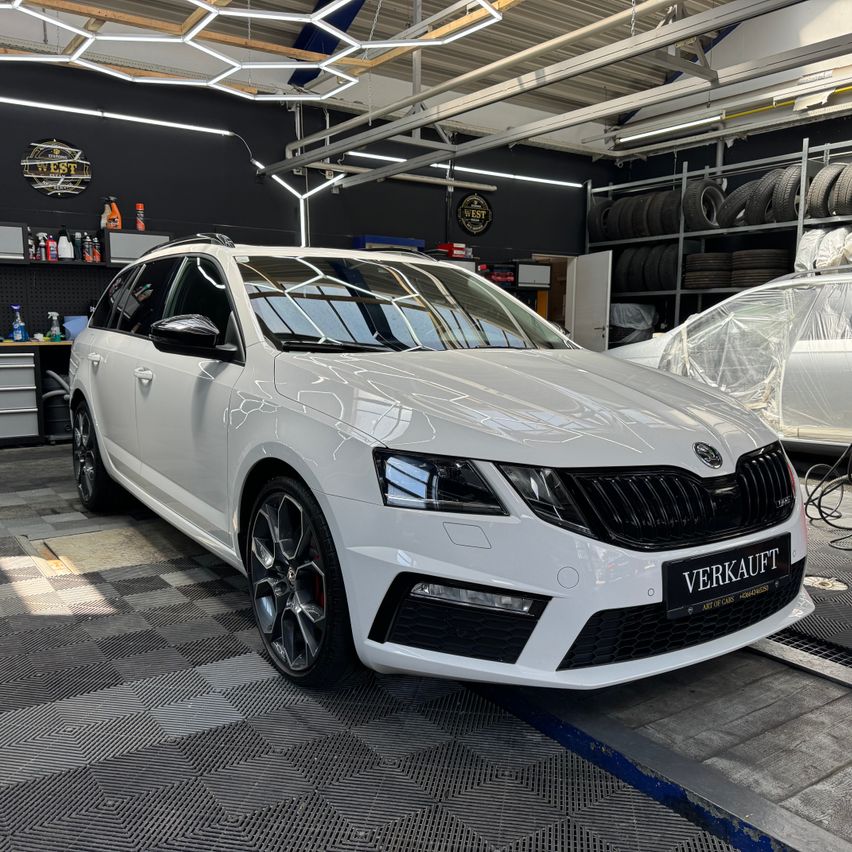 A white Skoda Octavia RS is parked in a garage with a blue floor and a white wall. The car has a black bumper, black rims, and a license plate that reads 'VERKAUFT'. There are tires and other items on the wall behind the car.