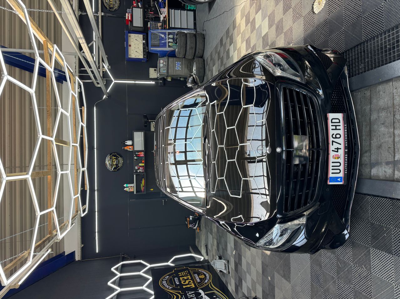 A black car with a hexagonal pattern on its hood is parked in a garage. The car's front bumper and grille are visible, and the license plate reads 'UU 176 HD'.