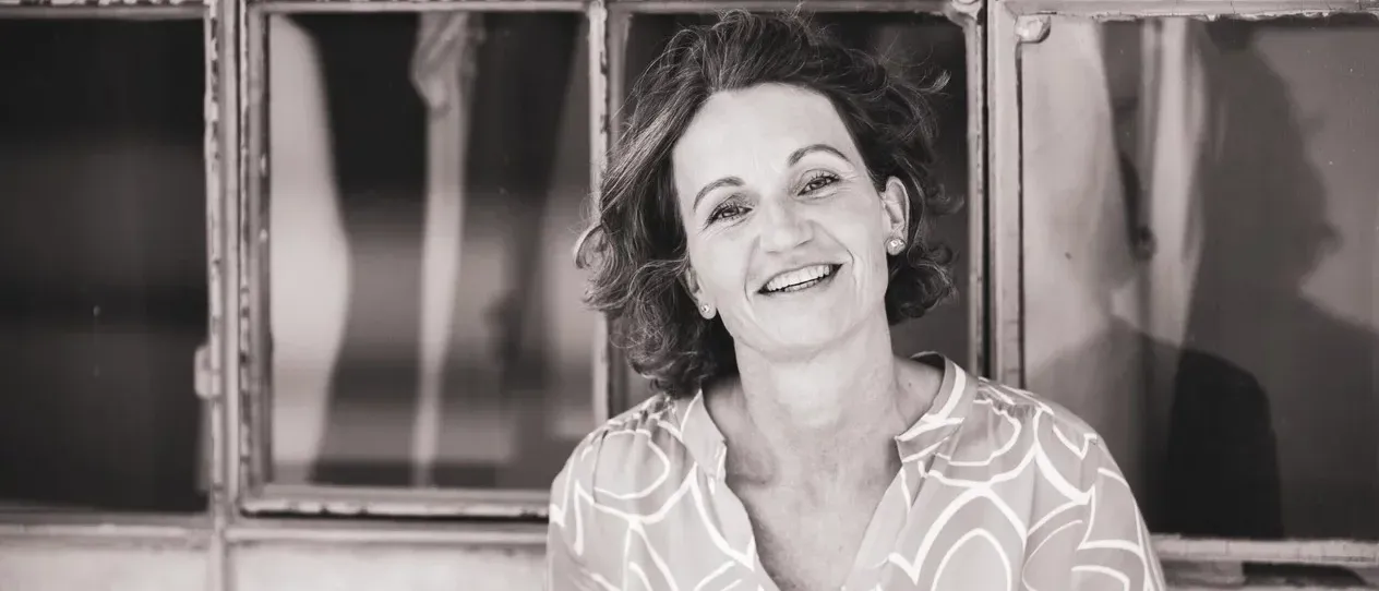 Ein Schwarz-Weiß-Porträt einer Frau mit kurzen, lockigen Haaren, die lächelt. Sie trägt ein gemustertes Hemd mit Kragen und Ohrringe. Im Hintergrund ist ein Glasfenster zu sehen.