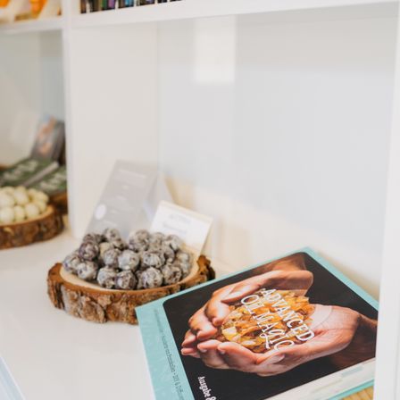Ein weißer Regal zeigt ein Buch mit dem Titel 'Fortgeschrittene Orakelmagie', das von einer Hand mit einem Kristall gehalten wird. In der Nähe steht ein hölzerner Teller mit runden, schwarzen Süßigkeiten. Der Hintergrund zeigt andere Regale mit verschiedenen Artikeln.