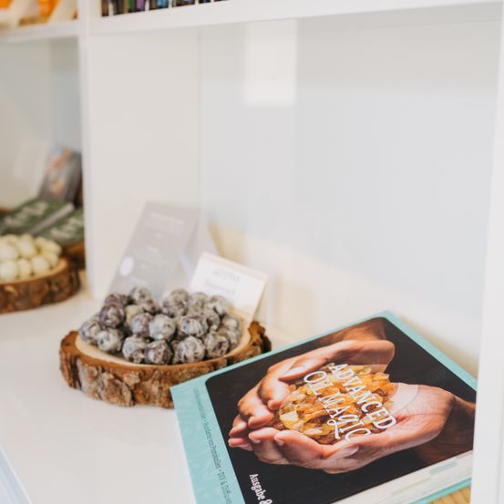Ein weißer Regal zeigt ein Buch mit dem Titel 'Fortgeschrittene Orakelmagie', das von einer Hand mit einem Kristall gehalten wird. In der Nähe steht ein hölzerner Teller mit runden, schwarzen Süßigkeiten. Der Hintergrund zeigt andere Regale mit verschiedenen Artikeln.