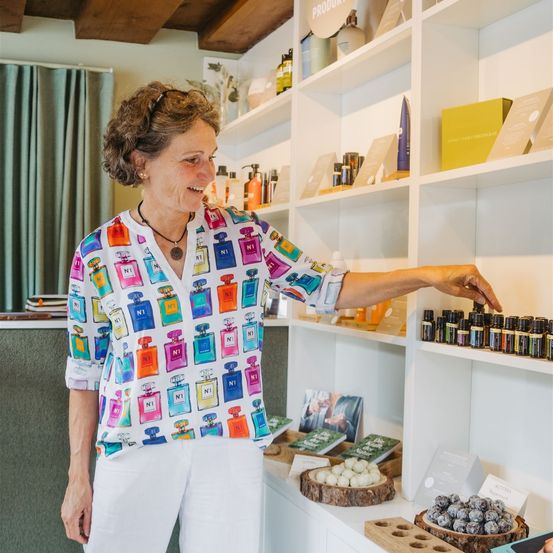 Eine lächelnde Frau in einem farbenfrohen Shirt zeigt auf Produkte in einem weißen Regal in einem Geschäft. Das Regal enthält verschiedene Flaschen und Artikel.