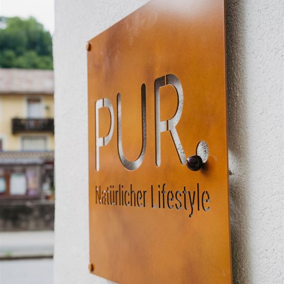 Ein braunes Schild an einer Wand zeigt in großen silbernen Buchstaben 'PUR'. Darunter steht 'Naturlicher Lifestyle'. Das Schild ist mit einem Metallring befestigt. Im Hintergrund ist ein verschwommenes Gebäude zu sehen.