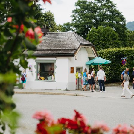 Ein weißes Haus mit einem grünen Regenschirm steht an der Ecke einer Straße, an der Menschen vorbeigehen. Bäume und Büsche umgeben das Gebiet.