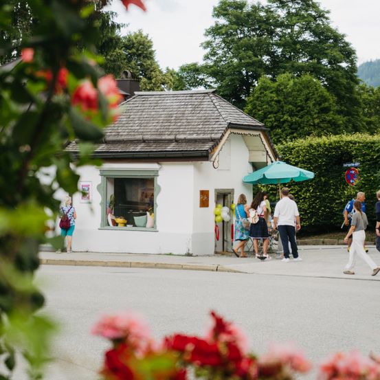 Ein weißes Haus mit einem grünen Regenschirm steht an der Ecke einer Straße, an der Menschen vorbeigehen. Bäume und Büsche umgeben das Gebiet.
