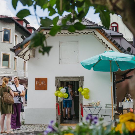 Zwei Frauen stehen vor einem weißen Gebäude mit der Aufschrift 'PUR.'. Eine Person mit Rucksack steht in der Tür. Draußen befindet sich ein türkisfarbener Schirm und Luftballons.