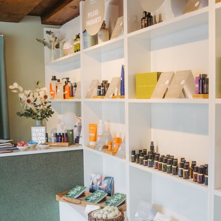 Ein Raum mit weißen Regalen, gefüllt mit verschiedenen Flaschen und Produkten. Ein Tisch hat einen Blumenstrauß, Bücher und Süßigkeiten. Ein Schild auf dem Tisch steht 'This Girl is on Fire'.