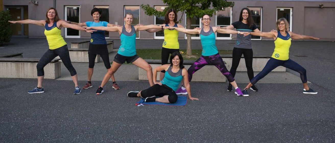 Eine Gruppe von Frauen in Trainingskleidung macht Yoga-Posen vor einem Gebäude mit einem Baum und Bänken.