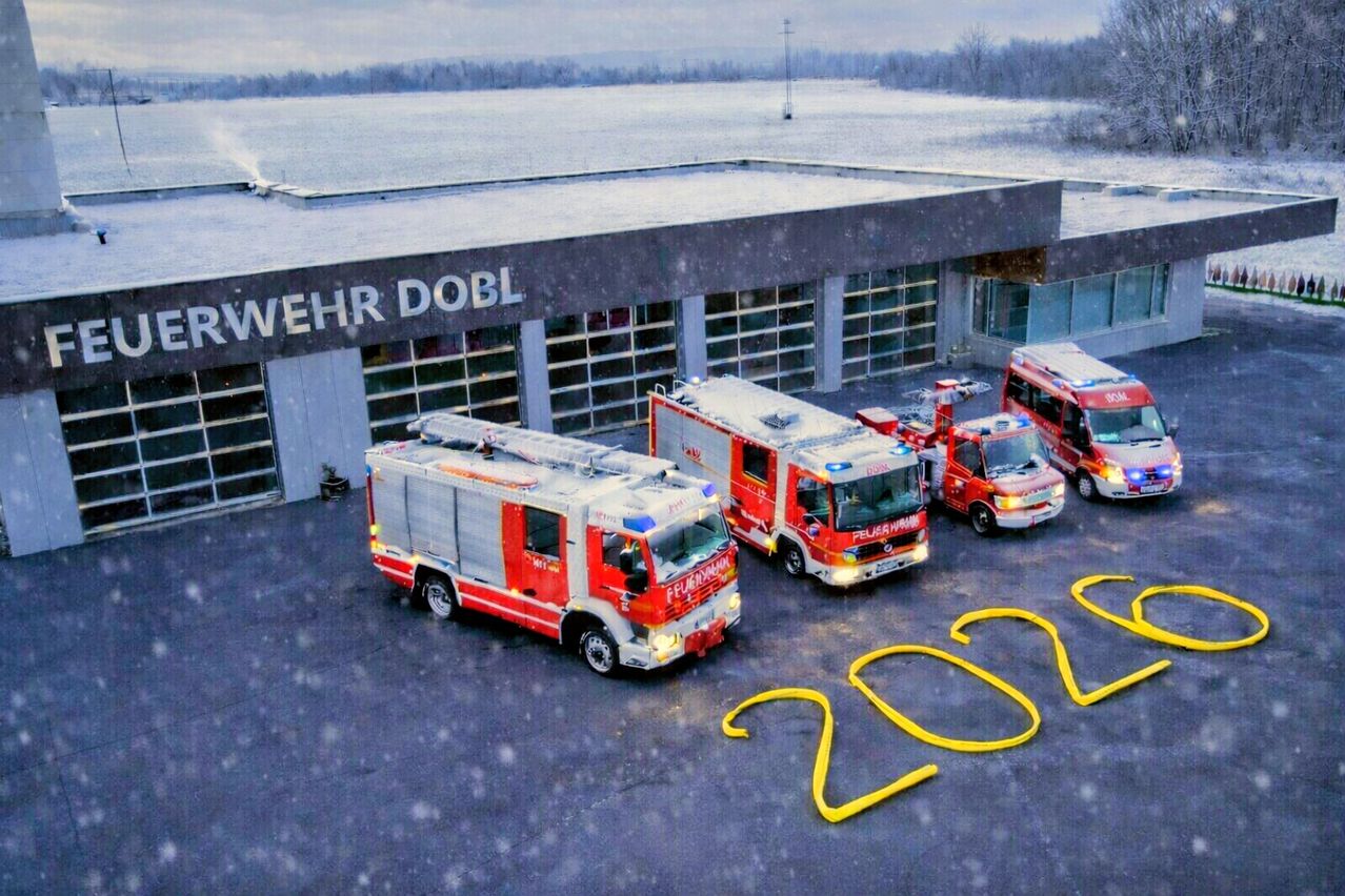 Mehrere Feuerwehrfahrzeuge sind vor einem Gebäude geparkt. Der Text 'wehr dobl' ist auf dem Gebäude zu sehen. Die Nummer 2021 ist auf der Straße vor den Fahrzeugen gemalt. Schnee fällt auf den Boden.