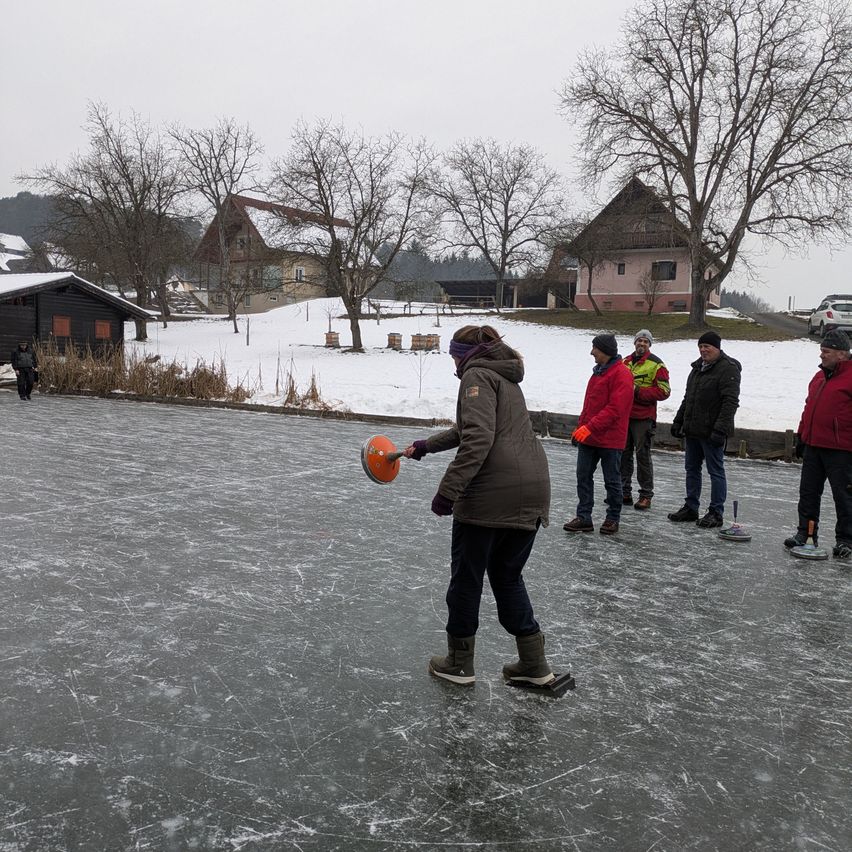 Eine Gruppe von Menschen, wahrscheinlich Freunde, spielt Eiscurling auf einem gefrorenen Teich, umgeben von einer verschneiten Landschaft mit Häusern im Hintergrund.