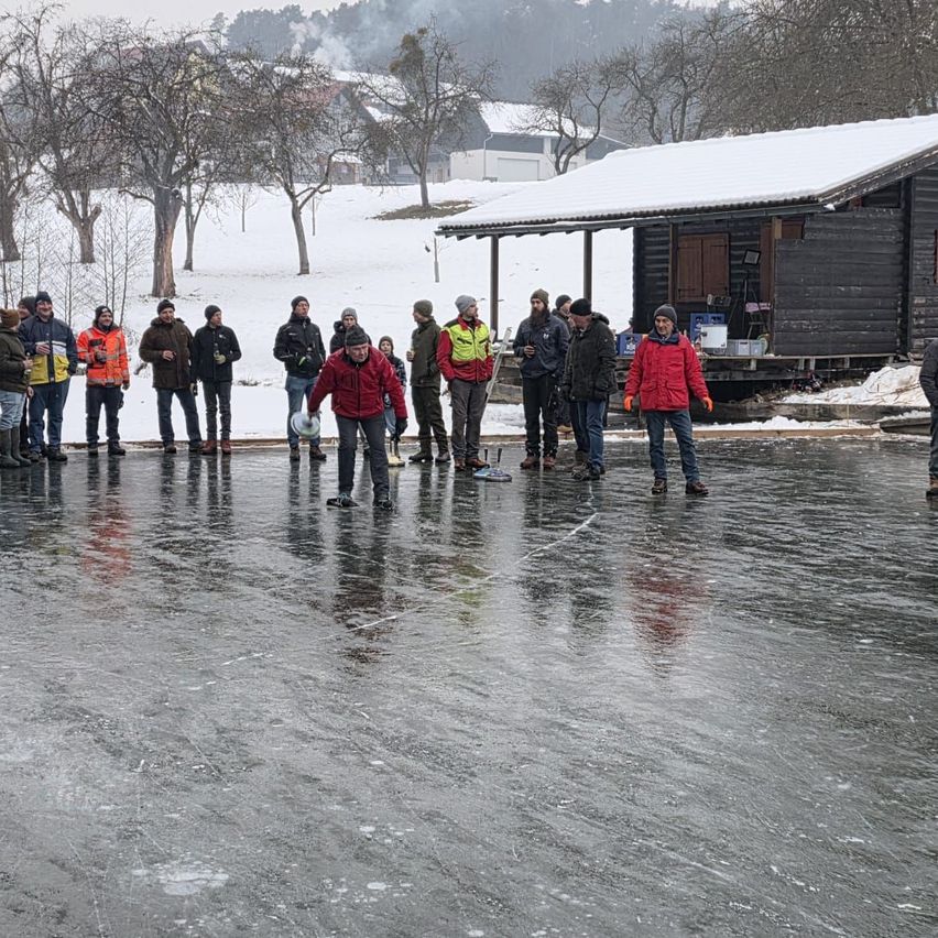 Eine Gruppe von Menschen steht auf einer Eisbahn in einer verschneiten Landschaft, mit einer Holzhütte im Hintergrund.