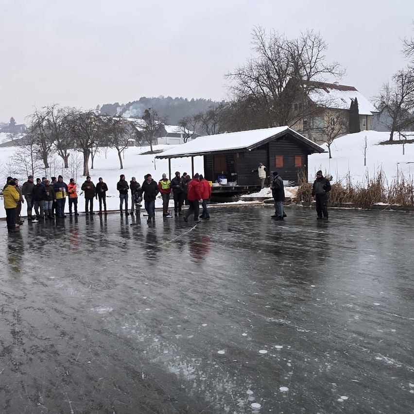 Eine Gruppe von Menschen versammelt sich auf einem gefrorenen See. Hinter ihnen steht eine kleine Hütte mit einem verschneiten Dach. Es gibt Bäume und Büsche in der Umgebung.