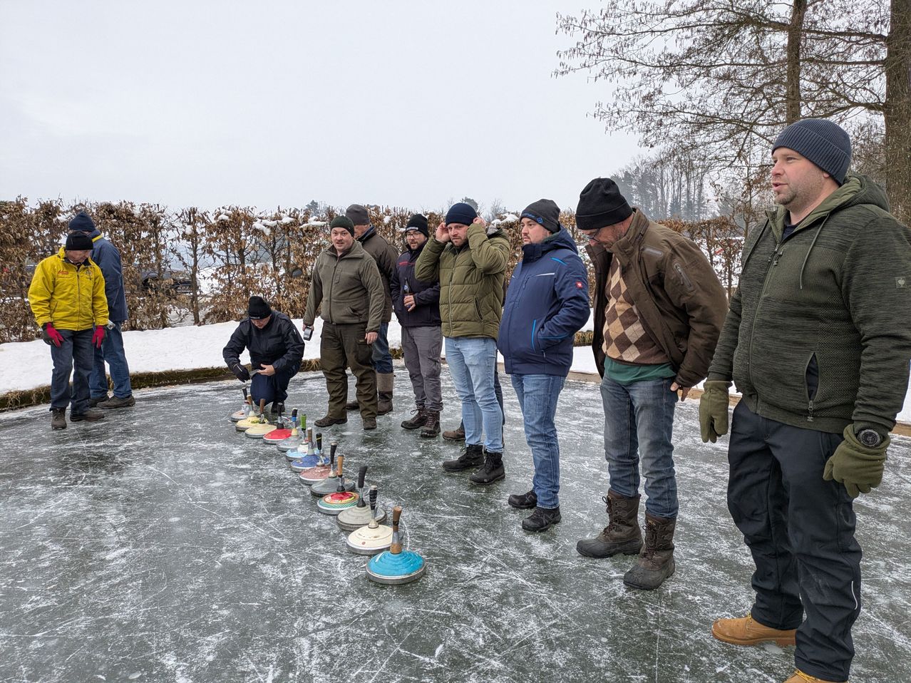 Eine Gruppe von Männern steht auf einer vereisten Oberfläche mit in einer Reihe angeordneten Curlingsteinen, möglicherweise in Vorbereitung auf ein Spiel.