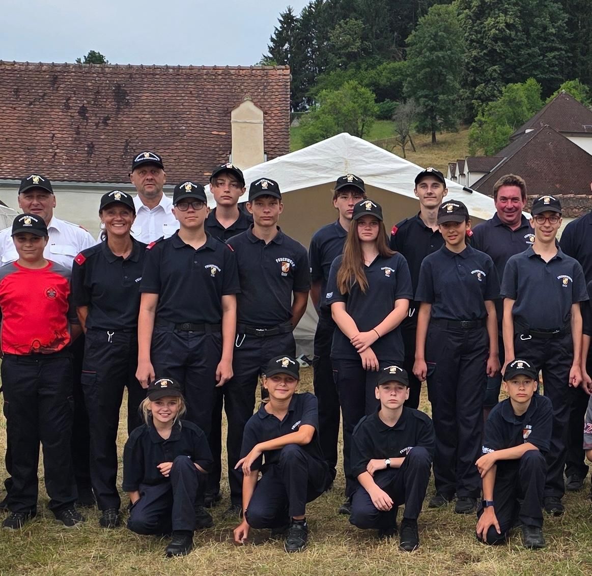 A group of uniformed individuals, possibly firefighters, pose for a photo in front of a building with a white tent in the background.