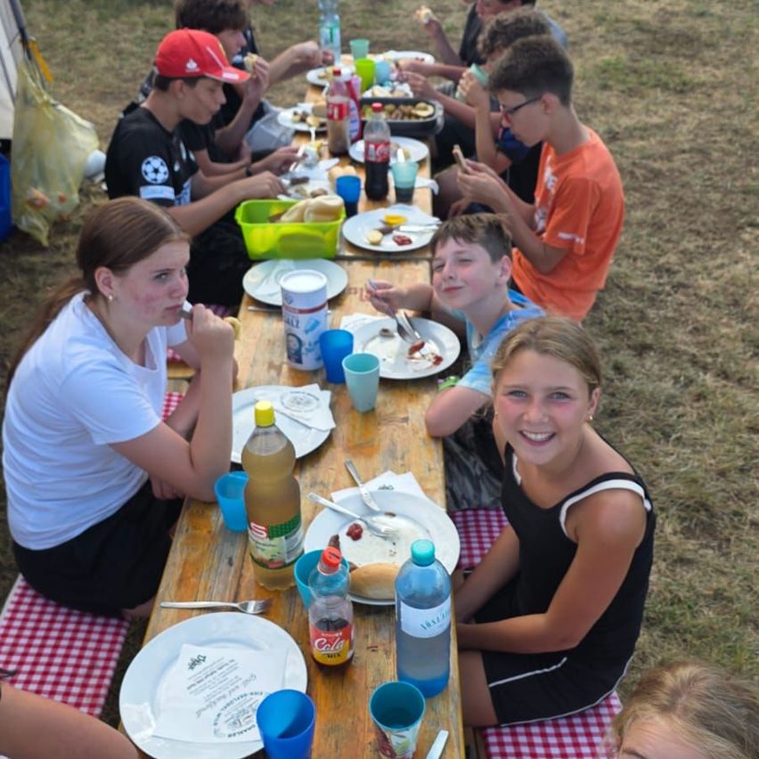 A group of children and young adults are sitting at a long picnic table, eating and drinking. The table is set with plates, cups, and various food items. The setting appears to be outdoors, with grass visible in the background.