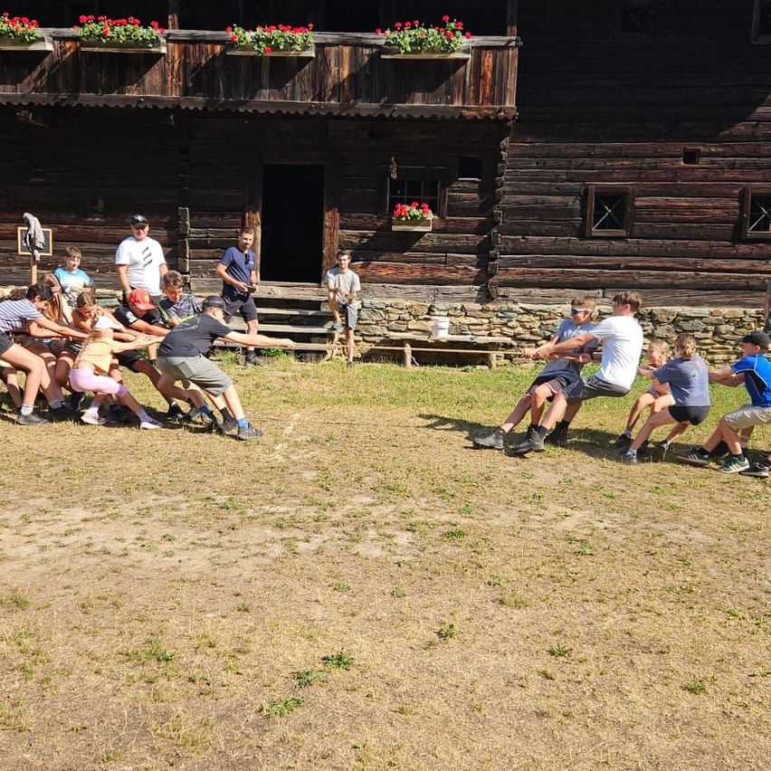A group of people are participating in a tug-of-war game in front of a wooden building with flower boxes on the balcony.