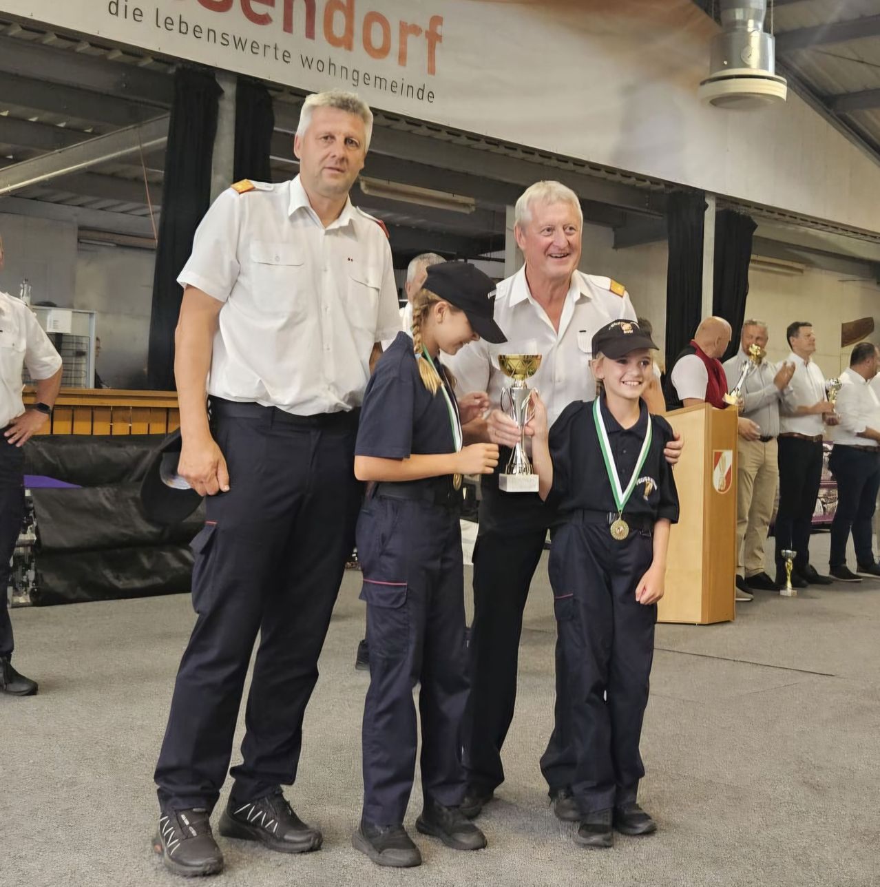 Zwei Männer in Uniformen stehen neben zwei Kindern, die Trophäen halten und lächeln. Hinter ihnen stehen mehrere Leute, einige klatschen. Ein Banner ist im Hintergrund.