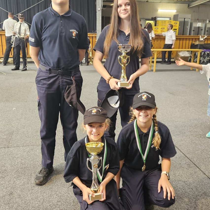 Drei Personen in Uniform, zwei Mädchen und ein Junge, posieren mit ihren Trophäen. Sie lächeln und knien auf dem Boden. Dahinter stehen zwei Personen mit den Händen auf den Hüften.