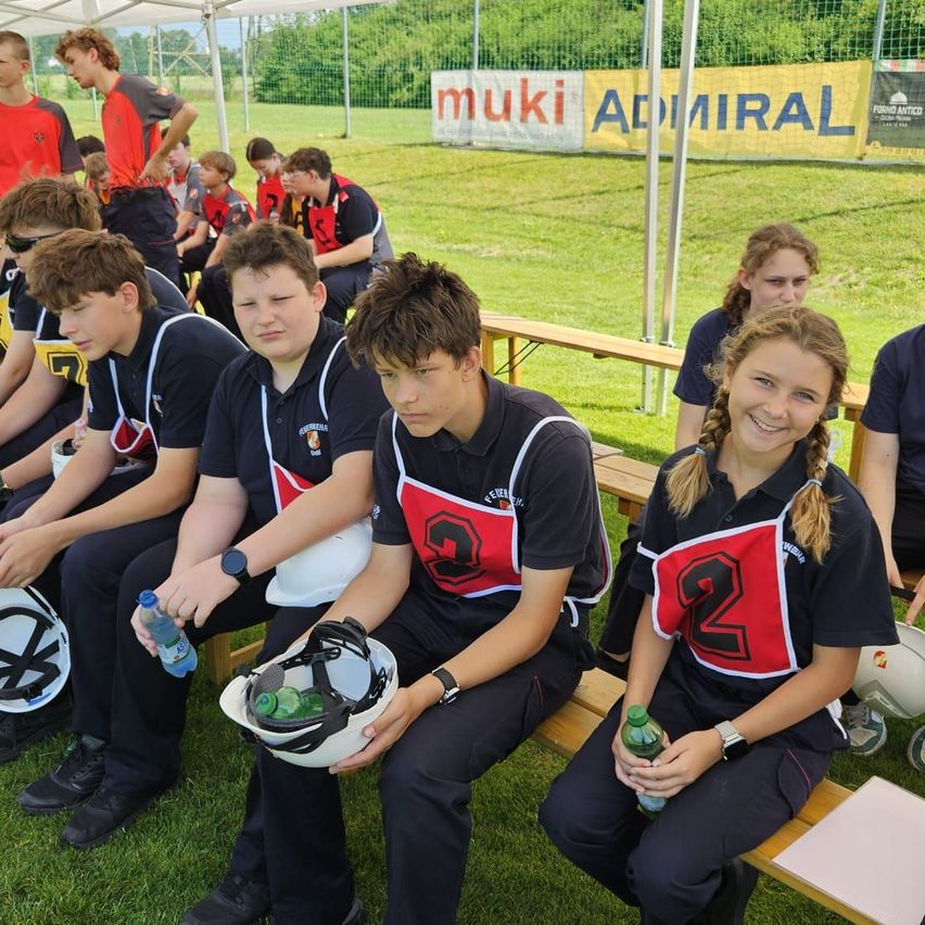 Eine Gruppe von Schülern in einheitlichen Uniformen sitzt unter einem Zelt auf einem Grasfeld, einige mit Helmen und Getränken. Im Hintergrund ist ein Banner mit der Aufschrift 'mukil ADMIRAL' zu sehen.