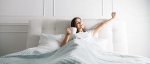 Eine Frau liegt lächelnd im Bett, hält eine Kaffeetasse und streckt ihre Arme.