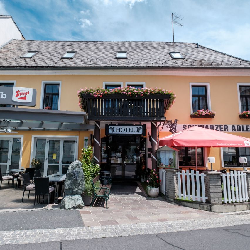 Die Vorderseite eines Hotels mit gelben Wänden, mehreren Fenstern und einem Balkon mit Blumen. Ein roter Schirm steht vor dem Eingang.