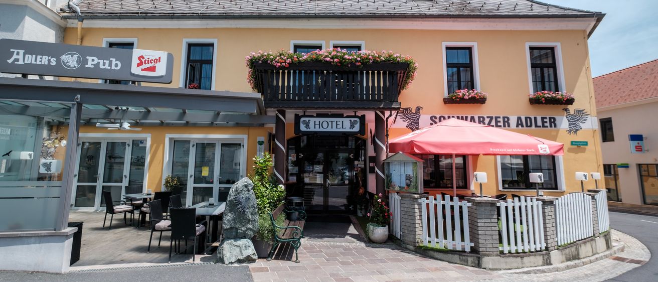 Die Vorderseite eines Hotels mit gelben Wänden, mehreren Fenstern und einem Balkon mit Blumen. Ein roter Schirm steht vor dem Eingang.