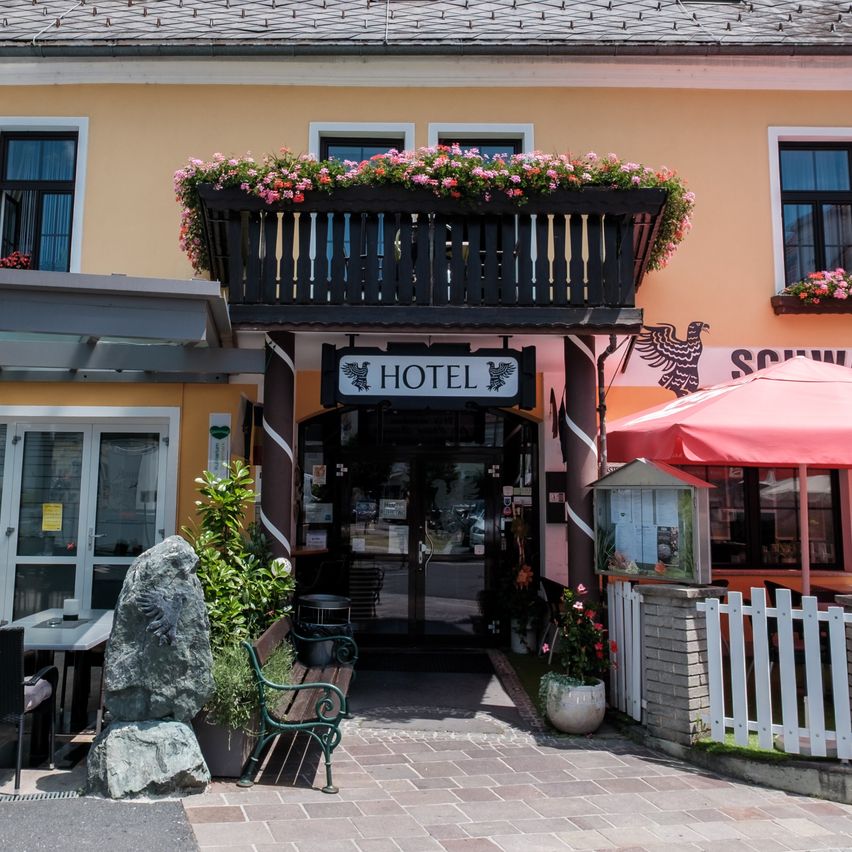 Die Vorderseite eines Hotels mit gelben Wänden, mehreren Fenstern und einem Balkon mit Blumen. Ein roter Schirm steht vor dem Eingang.