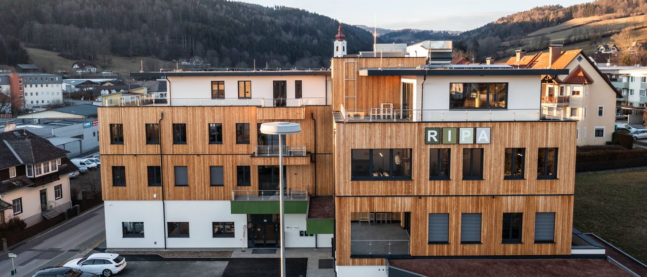 Ein Blick von oben auf das RIPA-Gebäude mit Holzfassaden, Glasfenstern und einem geneigten Dach. Es befindet sich in einem bergigen Gebiet mit üppiger Vegetation.