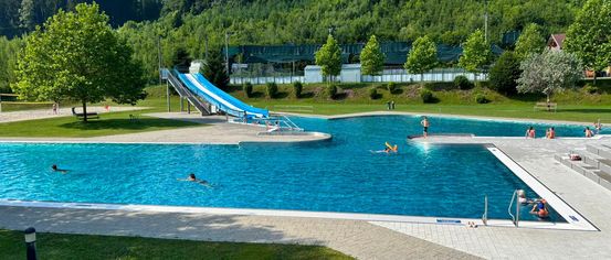 Ein öffentliches Schwimmbad mit einer Wasserrutsche ist von üppigem Grün und Bäumen umgeben. Mehrere Menschen schwimmen im Pool und andere entspannen sich am Rande.