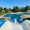 Freibad mit blauem Wasser, einer blauen Rutsche und schwimmenden Menschen. Bäume und Berge im Hintergrund.